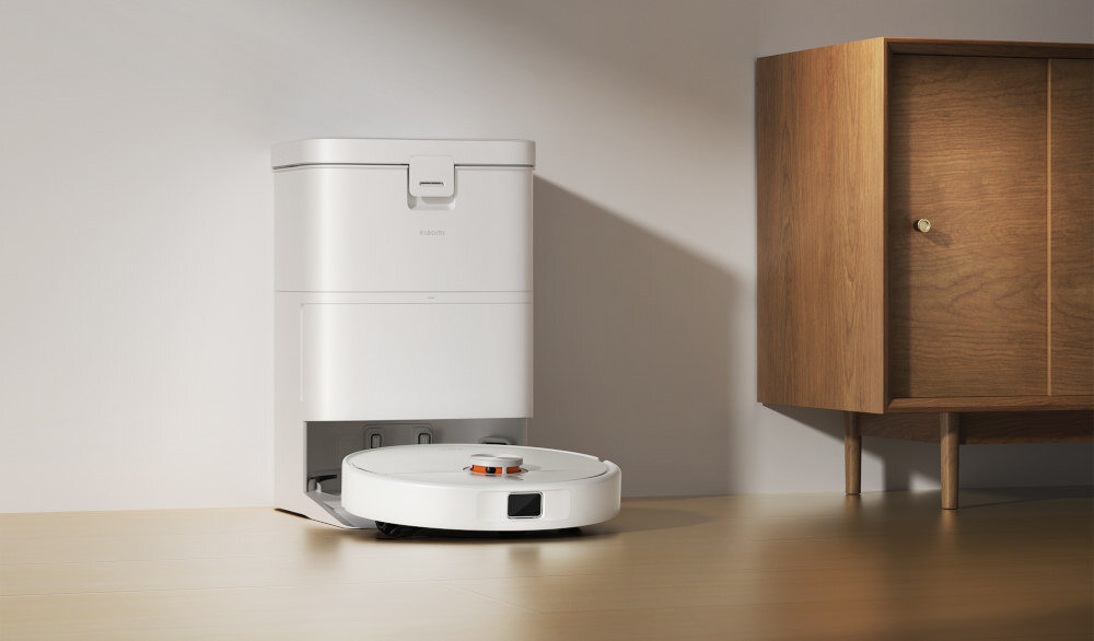 Na drewnianej powierzchni stoi biały robot sprzątający z widocznym logo \'XIAOMI\', obok stacji dokującej. W tle znajduje się drewniana szafka.
