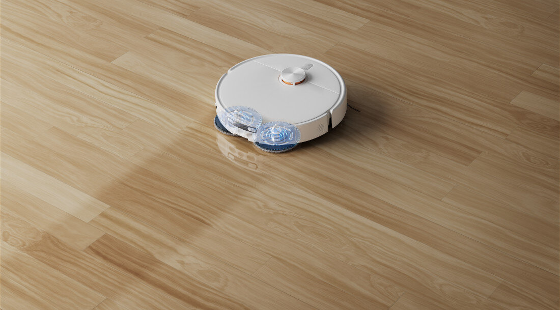 Na drewnianej podłodze znajduje się biały robot sprzątający o okrągłym kształcie. Robot ma widoczne niebieskie elementy z przodu.