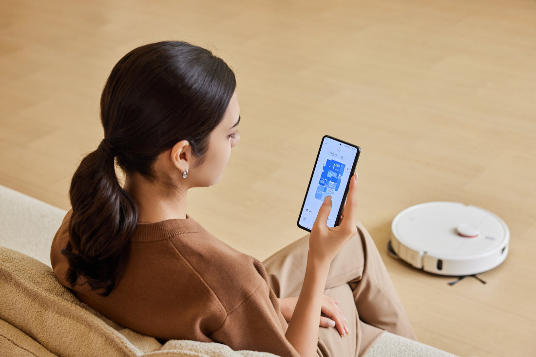 Osoba siedzi na kanapie, trzymając w ręku smartfon z wyświetloną aplikacją. Obok na podłodze znajduje się biały robot sprzątający.