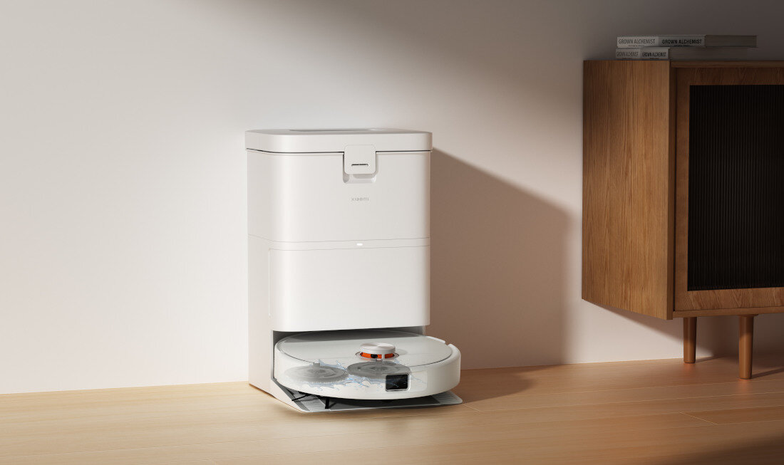 Na drewnianej podłodze stoi biały robot sprzątający z okrągłą podstawą, umieszczony w stacji dokującej. Obok znajduje się drewniana szafka z książkami na górze.
