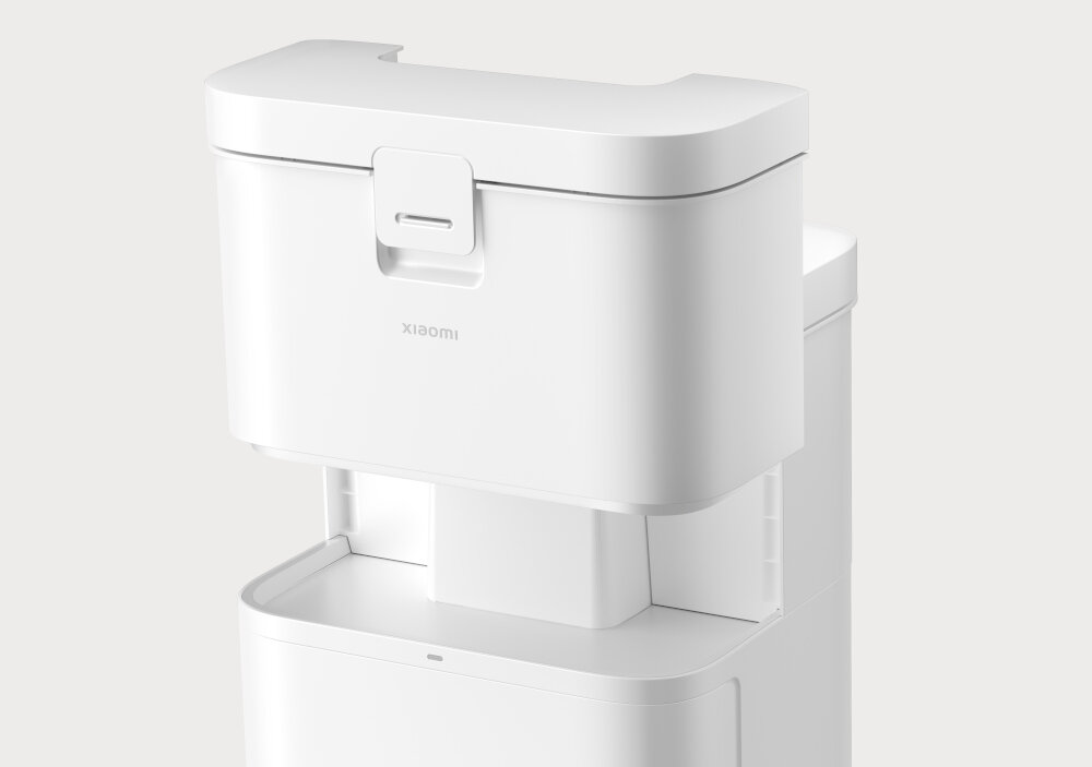 Biały pojemnik z logo \'xiaomi\' na górnej części, z uchwytem na klapie. Pojemnik jest częścią większego urządzenia, widocznego w tle.