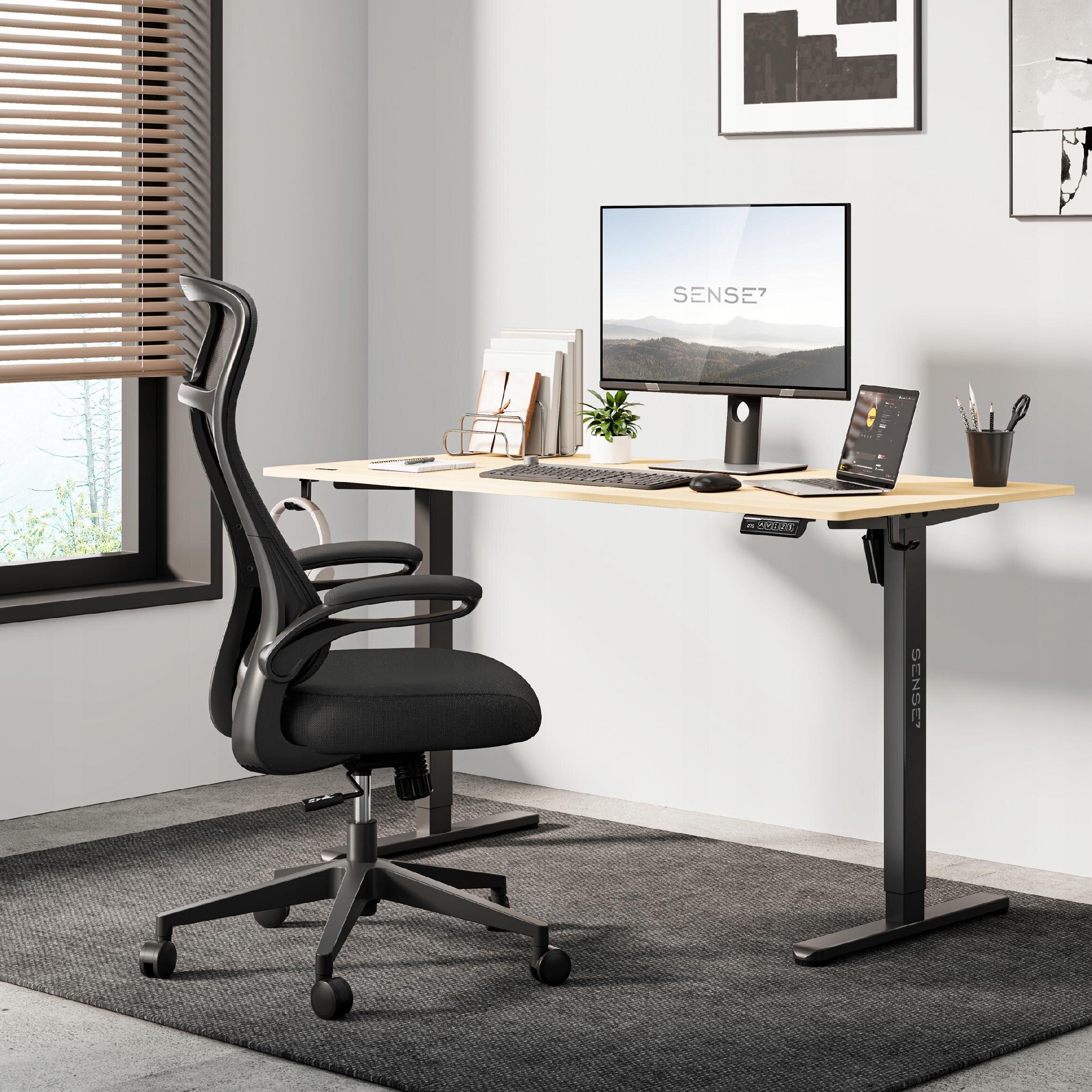 Na obrazie znajduje się czarny, ergonomiczny fotel biurowy przy biurku z monitorem, na którym widnieje napis \'SENSE7\'. W tle widać okno z żaluzjami oraz kilka dekoracji na ścianie.