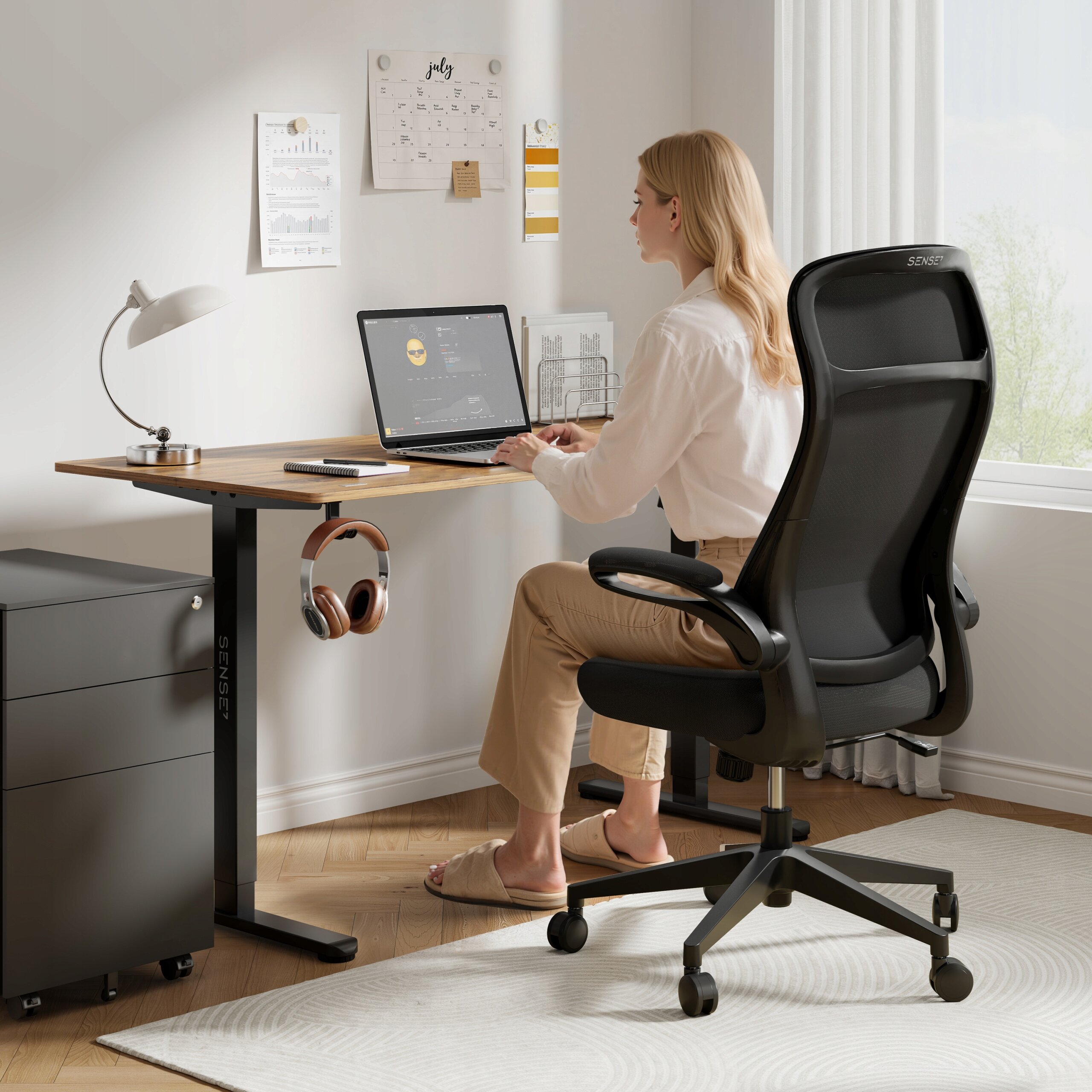Osoba siedzi przy biurku na czarnym, ergonomicznym fotelu biurowym SENSE7 Revo. Na biurku znajduje się laptop, lampka i słuchawki, a na ścianie wisi kalendarz i kilka notatek.