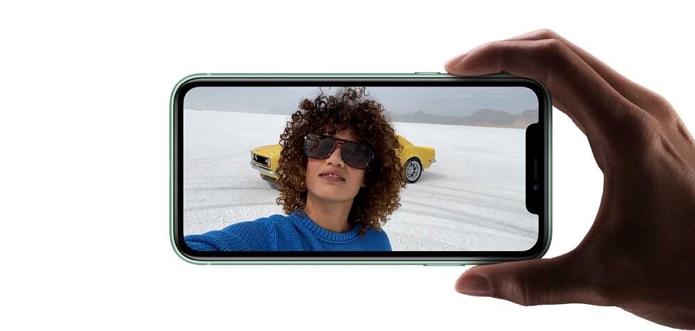 Osoba trzyma smartfon, na ekranie którego widać selfie kobiety w okularach przeciwsłonecznych z żółtym samochodem w tle. Smartfon ma cienkie ramki i zaokrąglone rogi.
