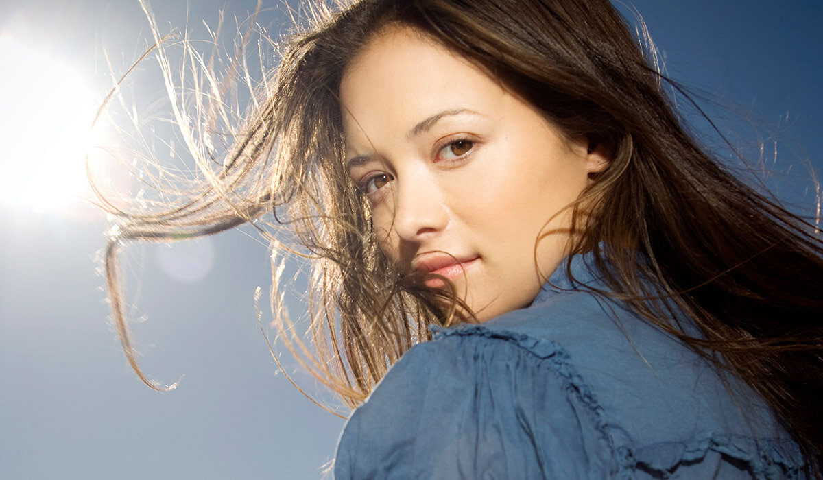 Osoba z długimi włosami patrzy w stronę aparatu, ubrana w niebieską bluzkę. W tle widać jasne niebo i słońce.