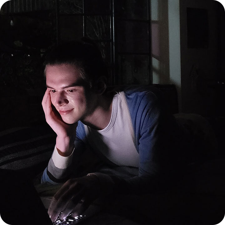 Osoba leży na łóżku w ciemnym pomieszczeniu, patrząc na ekran laptopa, który oświetla jej twarz.