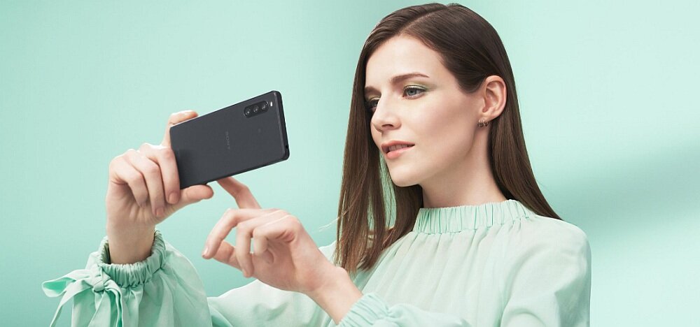 Osoba trzyma czarny smartfon, wykonując zdjęcie, na tle miętowej ściany. Smartfon ma trzy obiektywy aparatu z tyłu.