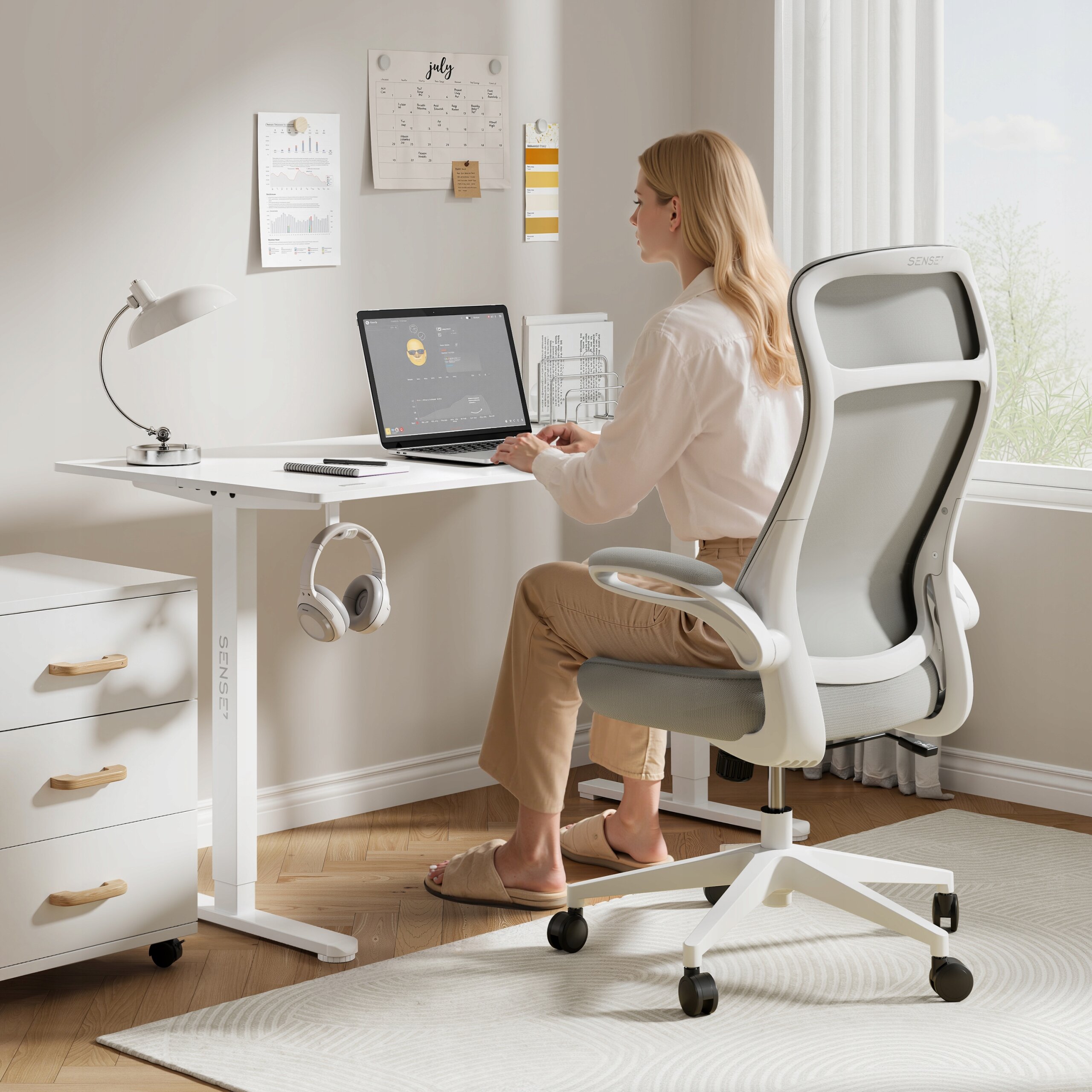 Osoba siedzi przy biurku na szarym, ergonomicznym krześle biurowym z regulacją. Na biurku znajduje się laptop, lampa i słuchawki, a na ścianie wisi kalendarz i notatki.