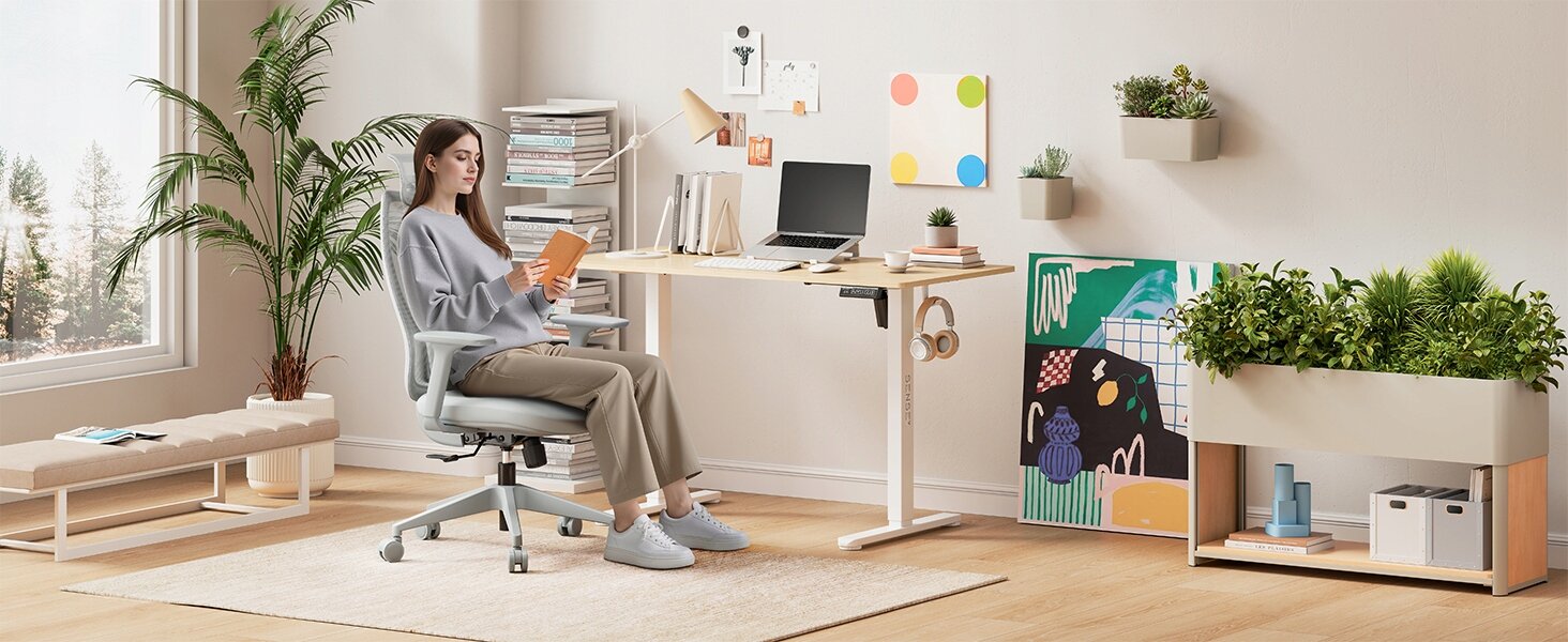 Osoba siedzi na szarym, ergonomicznym fotelu biurowym przy biurku w nowoczesnym, jasnym wnętrzu z roślinami i dekoracjami na ścianie. Na biurku znajduje się laptop, książki i inne akcesoria biurowe.