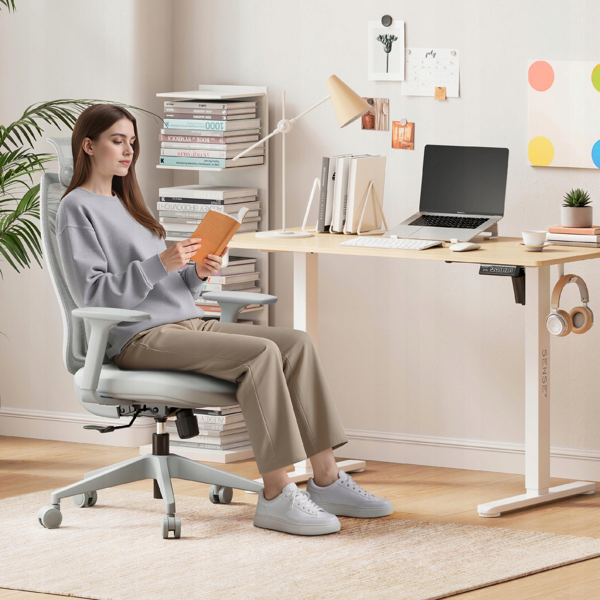 Osoba siedzi na szarym, ergonomicznym fotelu biurowym przy biurku, na którym znajduje się laptop, książki i lampka. W tle widać regał z książkami oraz dekoracje na ścianie.