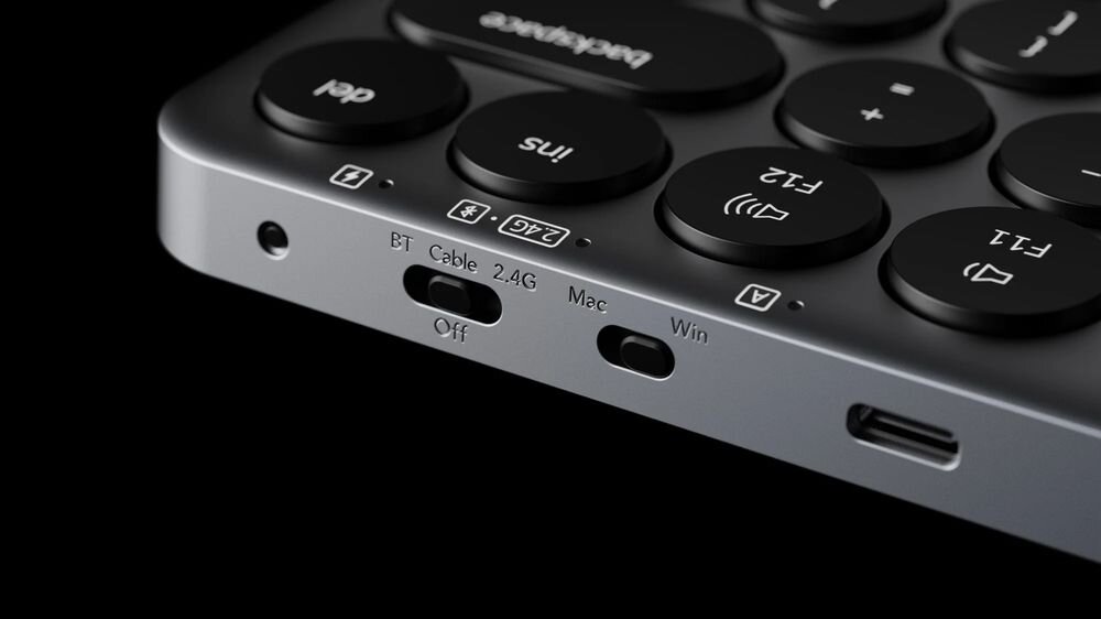 Zbliżenie na szarą klawiaturę z okrągłymi klawiszami i przełącznikami oznaczonymi jako BT, Cable, 2.4G, Mac, Win oraz portem USB-C. Widoczne jest logo \'keychron\' na jednym z klawiszy.
