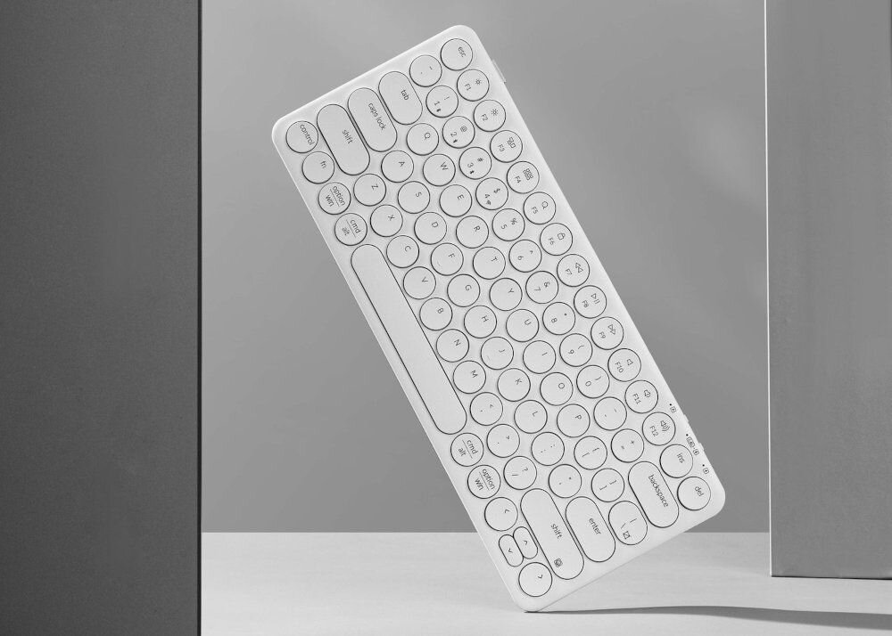 Na obrazie widoczna jest biała klawiatura z okrągłymi klawiszami, ustawiona pod kątem na szarym tle. Klawiatura ma minimalistyczny design i nie widać na niej żadnych napisów ani logo.