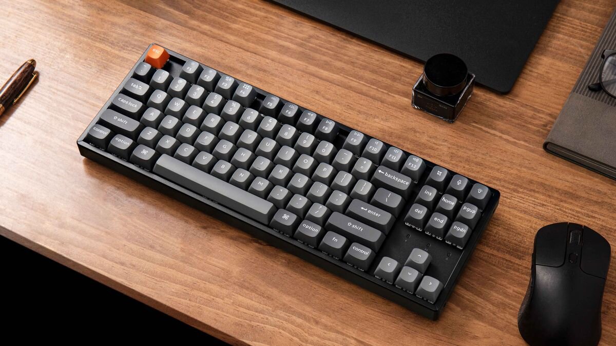 Keychron K8 Max Keychron K8 Max mechaniczna klawiatura bezprzewodowa solidną konstrukcję, trzy tryby łączności personalizację hot-swap i QMK/VIA - Klawiatura i mysz leżące na drewnianym biurku w otoczeniu artykułów piśmienniczych.