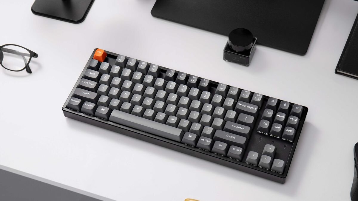 Keychron K8 Max Keychron K8 Max architektura oparta na kontrolerze ARM MCU 256 KB pamięci Flash częstotliwości odświeżania 1000 Hz trybie 2,4 GHz przewodowym - Klawiatura leżąca na biurku komputerowym w otoczeniu akcesoriów biurowych.