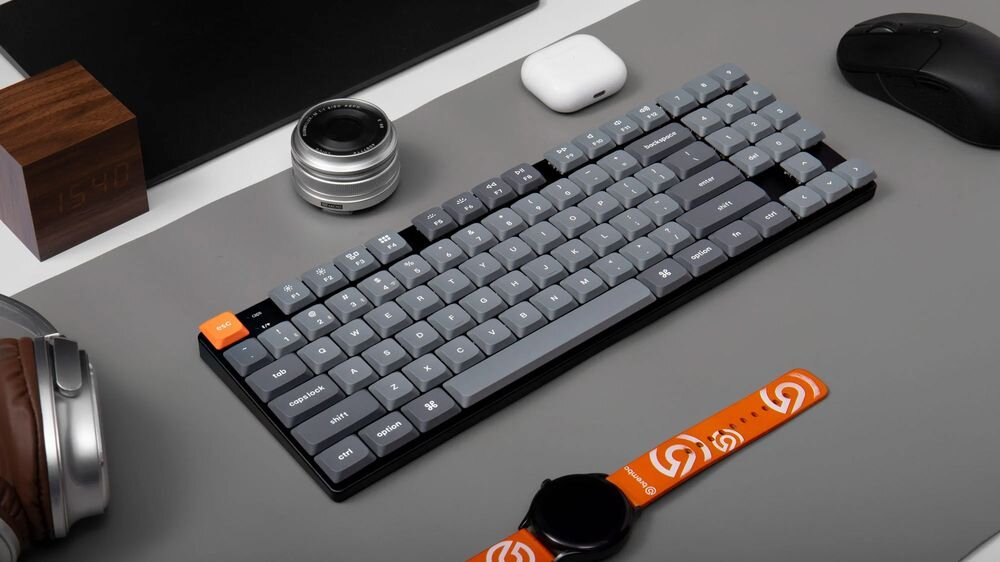 Keychron K13 Max niskoprofilowa klawiatura mechaniczna smukłą konstrukcję szerokimi możliwościami personalizacji wsparciu dla różnych trybów połączenia systemów macOS, Windows i Linux - klawiatura mechaniczna leżąca na ciemnoszarym biurku w otoczeniu gadżetów, takich jak smartwatch z pomarańczowym paskiem, słuchawki, obiektyw i mysz.
