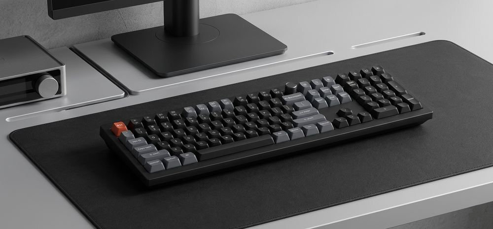 Na biurku znajduje się klawiatura mechaniczna z ciemnymi klawiszami i jednym wyróżniającym się czerwonym klawiszem. W tle widoczny jest monitor i inne elementy biurowe.