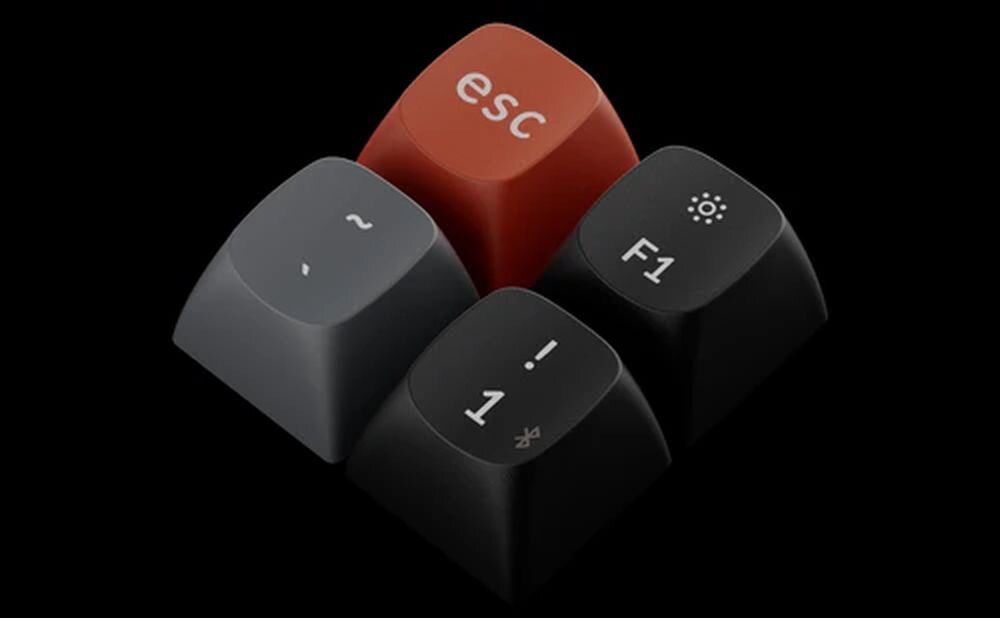 Na obrazie widoczne są cztery klawisze z klawiatury, w tym czerwony klawisz z napisem \'esc\'. Pozostałe klawisze mają symbole \'~\', \'F1\' z ikoną słońca oraz \'! 1\'.