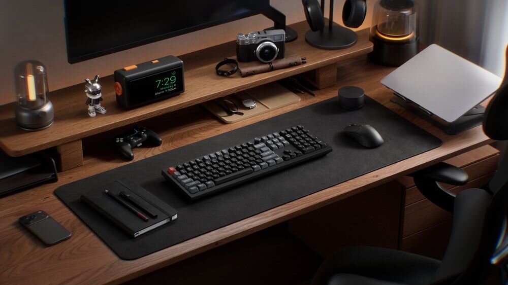 Na drewnianym biurku znajduje się klawiatura mechaniczna, mysz, laptop, aparat fotograficzny, zegar cyfrowy z wyświetlaczem 7:29 oraz kilka innych akcesoriów. Wszystko jest uporządkowane na dużej, ciemnej podkładce.