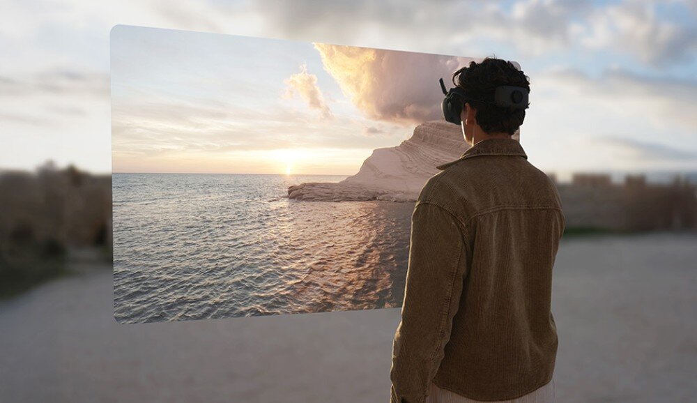 Osoba w goglach VR patrzy na wirtualny krajobraz przedstawiający morze i skały o zachodzie słońca. Tło jest rozmyte, co sugeruje skupienie na wirtualnym obrazie.