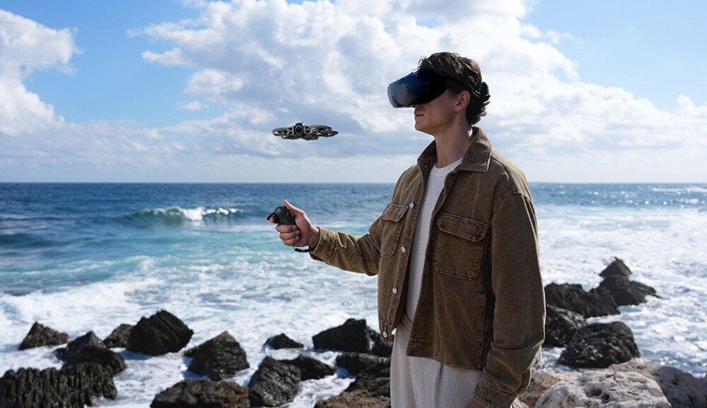 Osoba stoi na skalistym wybrzeżu, trzymając kontroler i nosząc gogle VR, podczas gdy w tle unosi się dron nad oceanem.
