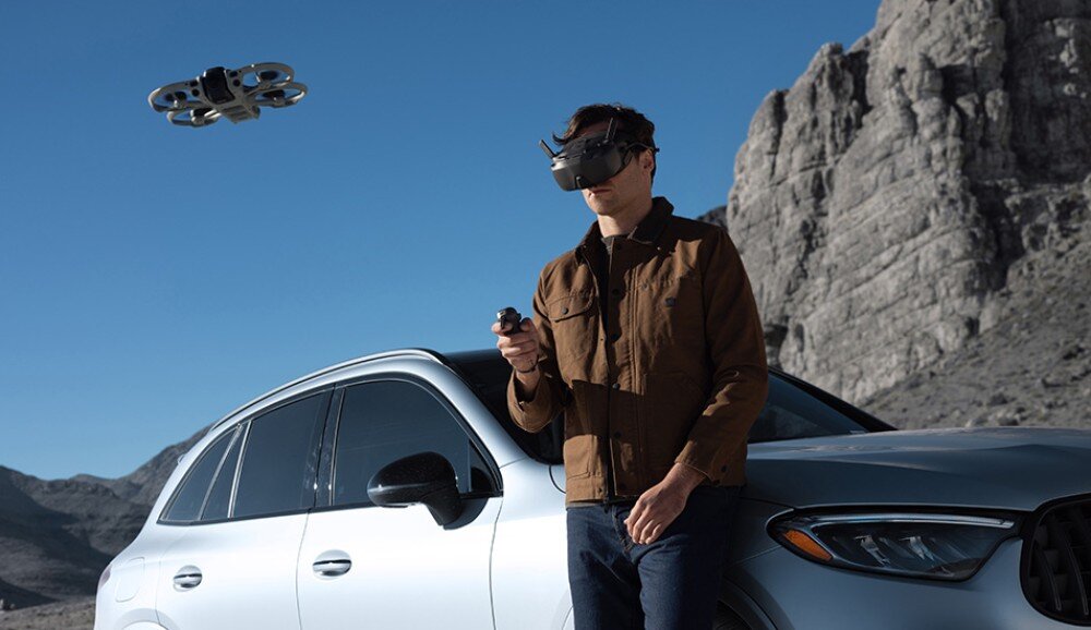 Osoba w goglach VR stoi obok srebrnego samochodu, trzymając kontroler, a nad nią unosi się dron. W tle widać skaliste góry i błękitne niebo.