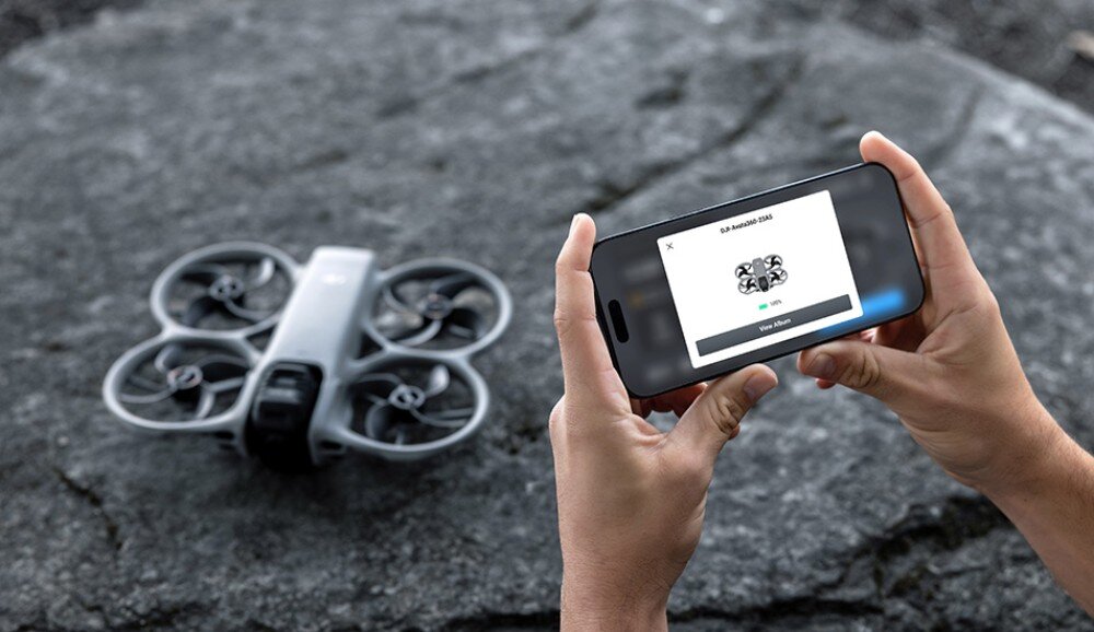 Osoba trzyma smartfon, na którym wyświetlany jest obraz drona z napisem \'DJI Avata 360\'. Obok na kamiennej powierzchni leży dron z wbudowaną osłoną śmigieł.