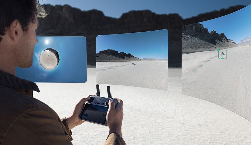 Osoba trzyma kontroler drona, patrząc na trzy wyświetlane obrazy przedstawiające pustynny krajobraz z górami. Na jednym z obrazów widoczny jest dron w locie.