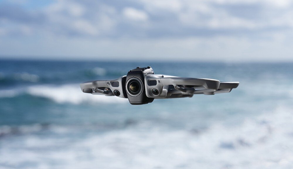 Dron unosi się w powietrzu na tle rozmytego krajobrazu morskiego. Widoczna jest jego kamera oraz elementy konstrukcyjne.