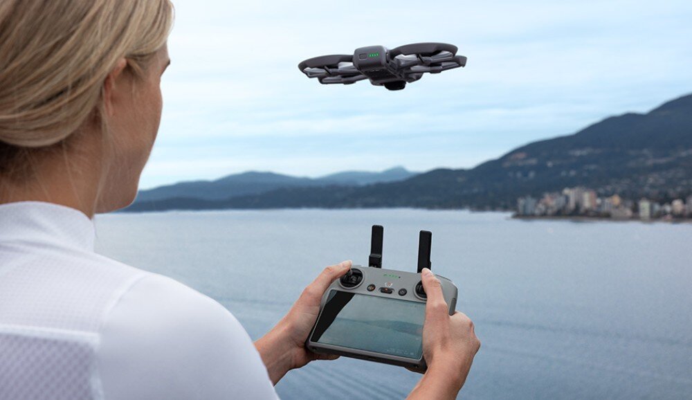 Osoba trzyma kontroler drona, patrząc na latający dron nad wodą z widokiem na góry w tle. Dron ma wbudowaną osłonę śmigieł.