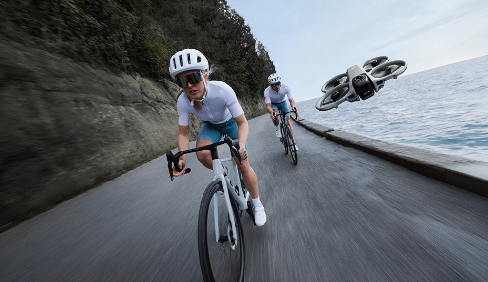 Dwie osoby na rowerach jadą wzdłuż drogi przy brzegu morza, a obok nich unosi się dron. Dron ma wbudowaną osłonę śmigieł i jest w kolorze szarym.