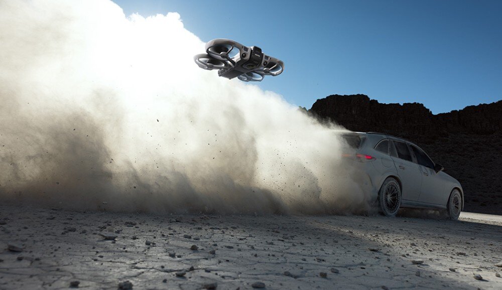 Dron unosi się nad ziemią, wzbijając chmurę kurzu obok jadącego samochodu na pustynnym terenie. W tle widać skaliste wzgórza i jasne niebo.