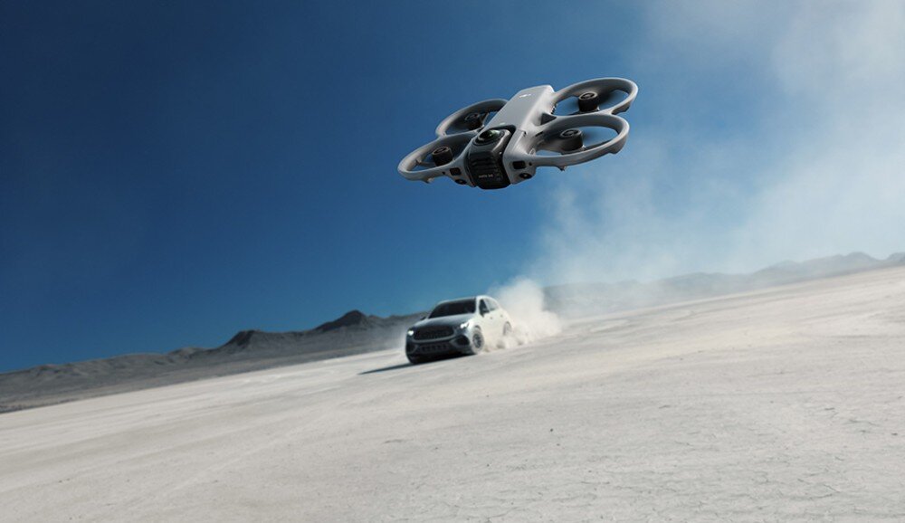 Na obrazie widać drona unoszącego się nad pustynnym terenem, w tle znajduje się samochód. Dron ma wbudowaną osłonę śmigieł i jest w kolorze jasnym.