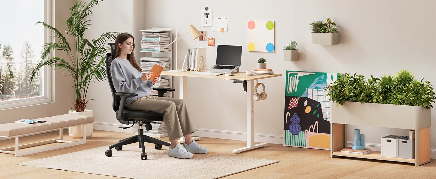 Osoba siedzi na czarnym, ergonomicznym fotelu biurowym przy biurku w nowoczesnym, jasnym wnętrzu z roślinami i dekoracjami na ścianie. Na biurku znajduje się laptop, książki i inne akcesoria biurowe.