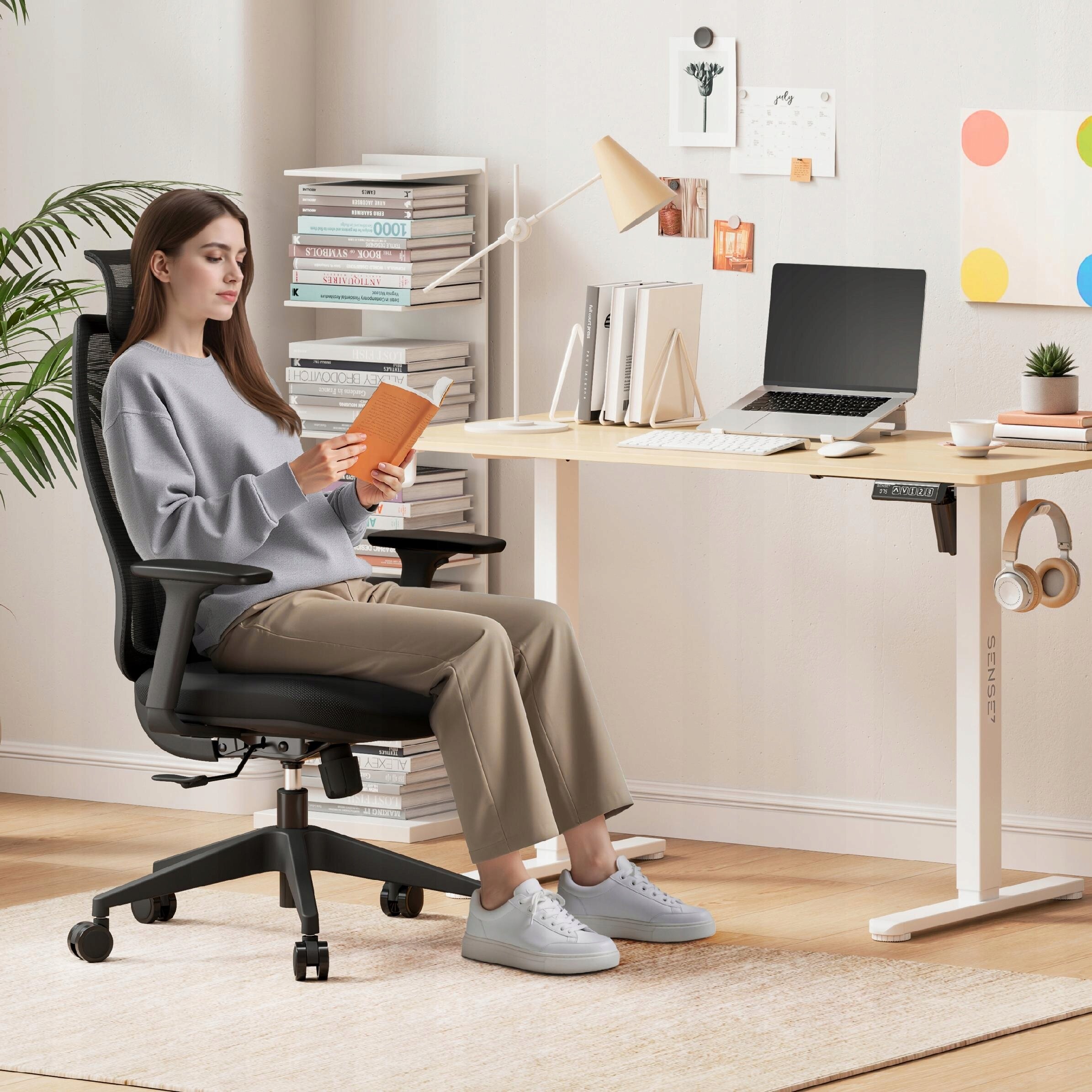 Osoba siedzi na czarnym, ergonomicznym krześle biurowym przy biurku z laptopem i książkami. Na biurku znajduje się lampa, a na ścianie widać kilka dekoracji i półkę z książkami.