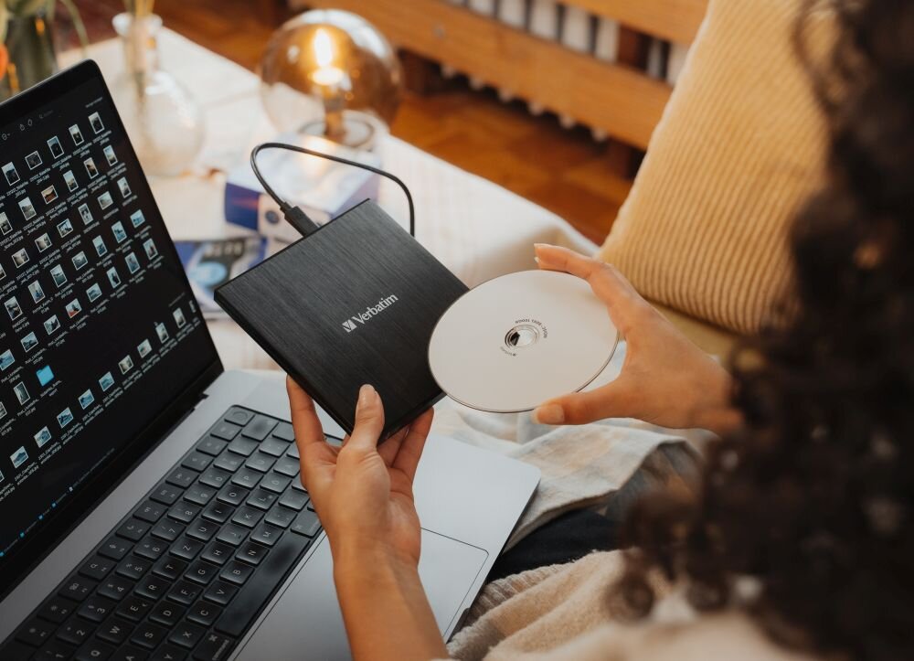 Napęd VERBATIM (43886) Osoba trzyma czarny napęd optyczny i biały krążek nad laptopem, na ekranie widoczne są miniatury plików zewnętrzna nagrywarka USB 3.2 Gen1