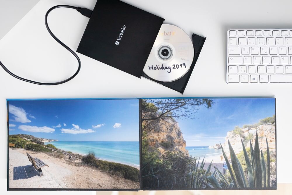 Napęd VERBATIM (43886) Czarny napęd optyczny z wysuniętą tacką, w środku znajduje się płyta z napisem 'Holiday 2019', obok otwarty album ze zdjęciami plaży i klifów oraz biała klawiatura zasilanie z USB-C, idealna do laptopów