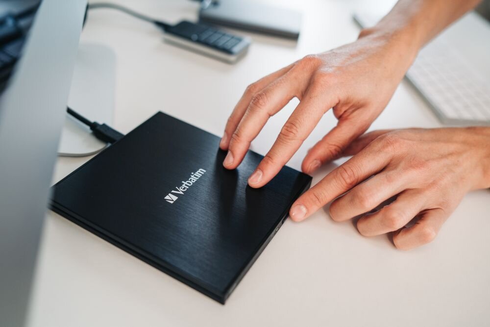 Napęd VERBATIM (43886) Zbliżenie na dłonie zamykające czarny napęd optyczny stojący na biurku obok klawiatury i innych urządzeń podłączonych do komputera archiwizacja danych na lata