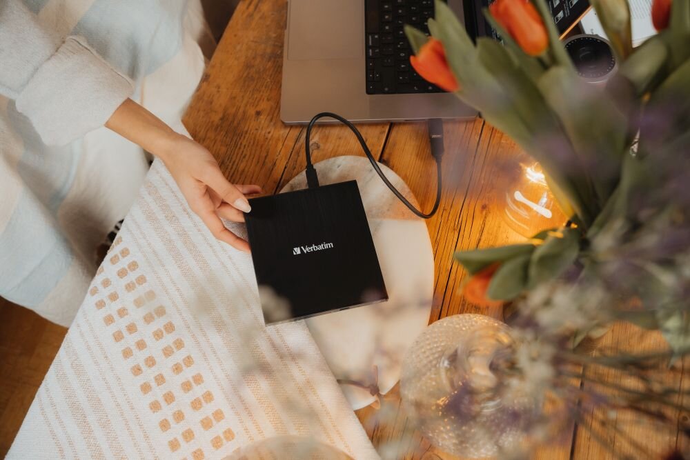 Napęd VERBATIM (43886) Osoba siedzi przy drewnianym stole, na którym stoi laptop, a obok leży czarny napęd optyczny, na pierwszym planie widać bukiet tulipanów i szklane naczynia Nagrywarka USB-C z kablem, adapterem i Nero Burn & Archive