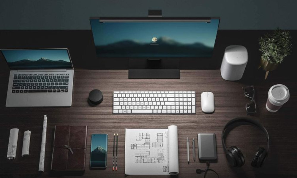 Na biurku znajduje się monitor z zamontowaną lampką, klawiatura, mysz, laptop, słuchawki, notatnik, kubek z napojem, roślina doniczkowa i inne akcesoria biurowe. Na ekranie monitora widoczny jest obraz górski z ikoną użytkownika pośrodku.