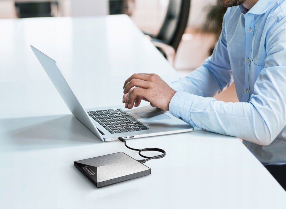 Napęd ZENWIRE SY881 Mężczyzna w jasnoniebieskiej koszuli pracuje przy laptopie na białym biurku. Obok laptopa leży podłączony napęd optyczny, przeznaczenie, zastosowanie, formaty płyt