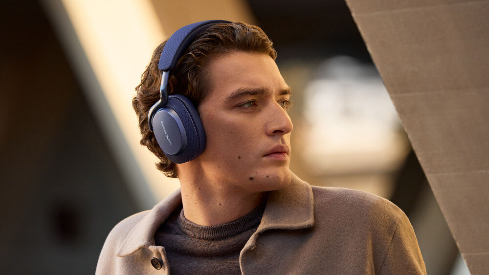 Osoba nosi czarne słuchawki nauszne z widocznym logo \'Bowers & Wilkins\'. W tle znajduje się rozmyty, nowoczesny budynek.