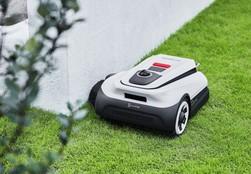 Robot koszący w kolorze białym z czarnymi elementami porusza się po trawie wzdłuż białej ściany. Na urządzeniu widoczne jest logo \'ECOVACS\'.