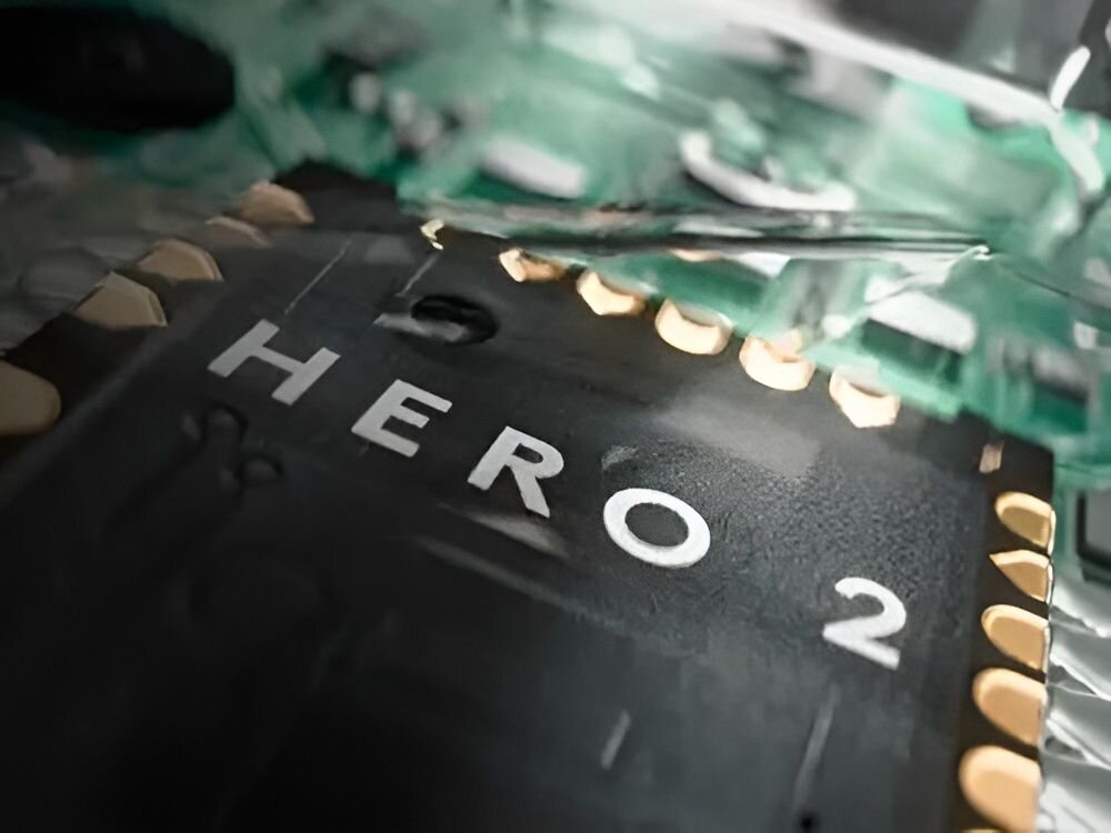 Zbliżenie na czarny układ scalony z napisem \'HERO 2\'. Wokół widoczne są złote styki oraz zielone elementy elektroniczne.