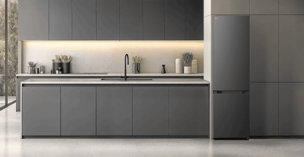 LODÓWKA LG GBBSJ2CCEP Minimalistyczna kuchnia w tonacji grafitowej z długą wyspą i gładkimi frontami bez uchwytów, oświetlona subtelną listwą LED pod szafkami wiszącymi. Po prawej stronie zabudowy stoi wolnostojąca lodówka w kolorze dopasowanym do mebli, tworząc spójną, elegancką aranżację