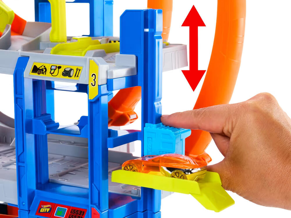 Tor Hot Wheels Mega Garaż Gigapętla + auto JBM74 Dwie dłonie trzymające czarny kontroler (pilot). Obok, na drewnianej podłodze, zdalnie sterowany samochód Hot Wheels Monster Truck Shark przewrócony na bok, pokazujący podwozie i duże opony z pomarańczowymi felgami.