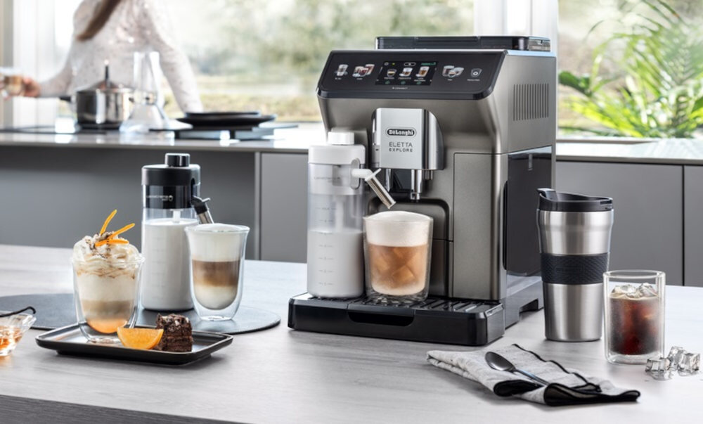 Na blacie kuchennym stoi ekspres do kawy z logo \'De\'Longhi Eletta Explore\'. Obok ekspresu znajdują się różne napoje kawowe w szklankach i kubkach oraz akcesoria do kawy.