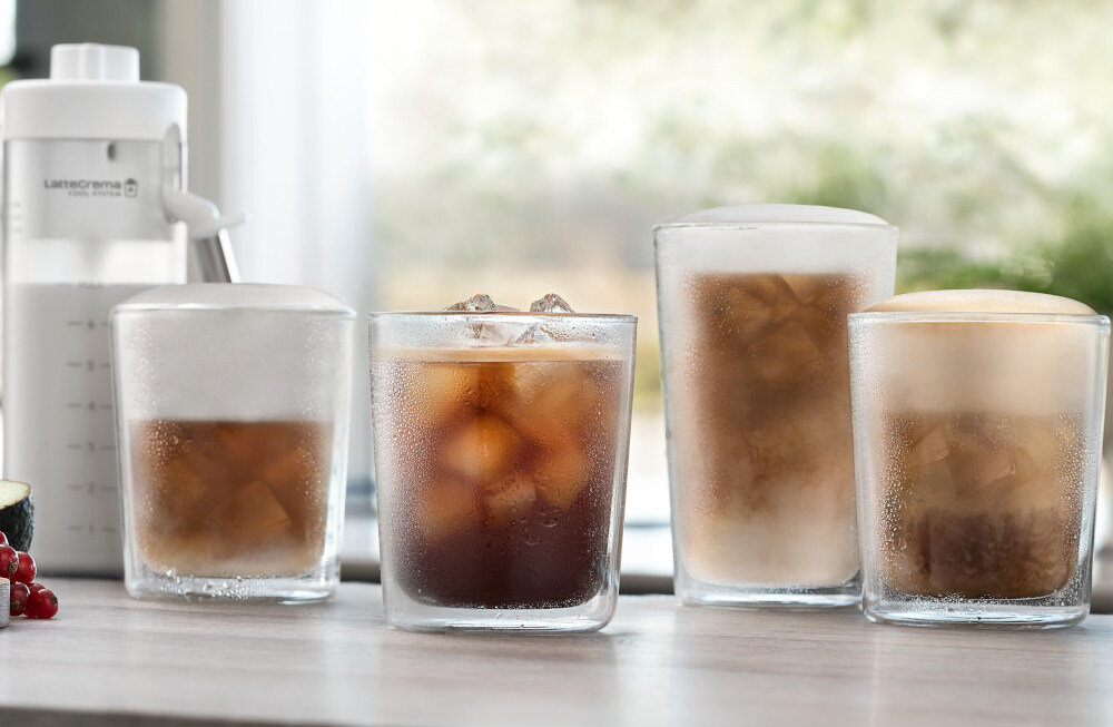 Na blacie stoją cztery szklanki z różnymi napojami, w tym kawa z lodem i mlekiem. Obok znajduje się biały pojemnik z napisem \'LatteCrema System\'.