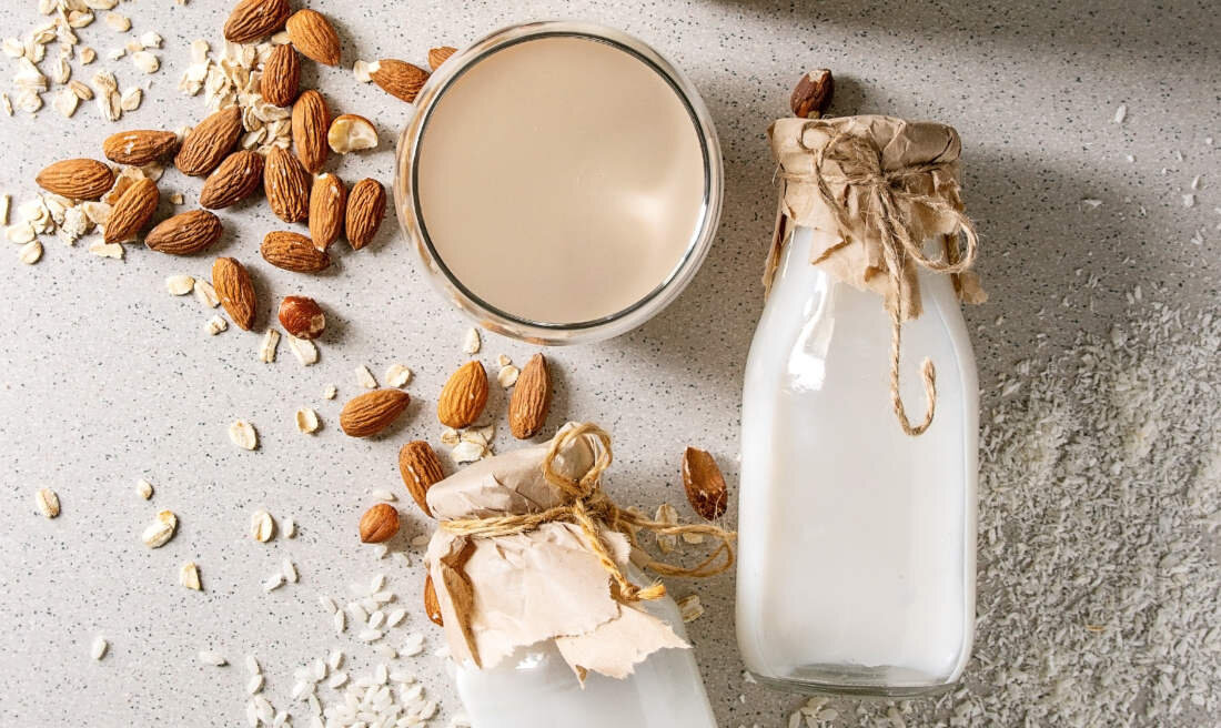 Na blacie znajdują się dwa szklane słoiki z białym płynem, zakryte papierem i przewiązane sznurkiem. Obok leży szklanka z jasnym napojem oraz rozsypane migdały i płatki owsiane.