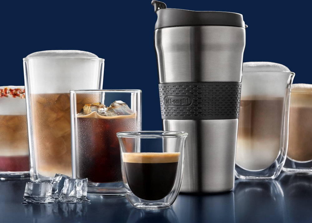 Na obrazie widoczne są różne szklanki z napojami kawowymi oraz metalowy kubek termiczny z logo \'De\'Longhi\'. W tle znajdują się kostki lodu i różnorodne kawy, w tym z mleczną pianką i na zimno.