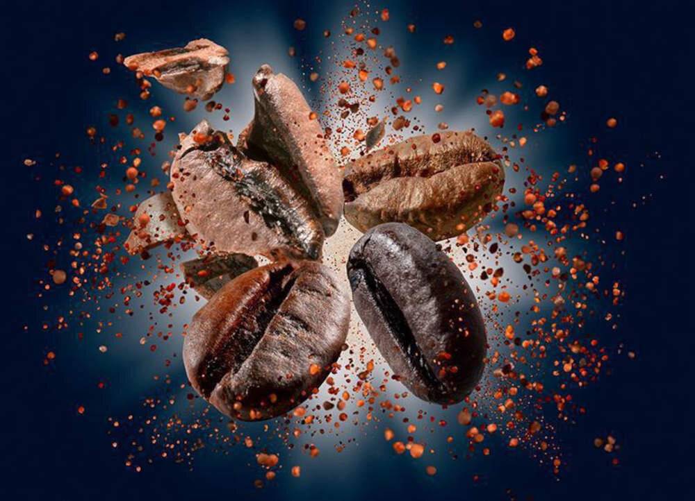 Na obrazie widoczne są unoszące się ziarna kawy otoczone drobnymi cząstkami kawy na ciemnym tle. Ziarna są w różnych odcieniach brązu, a wokół nich rozprzestrzeniają się drobinki kawy.